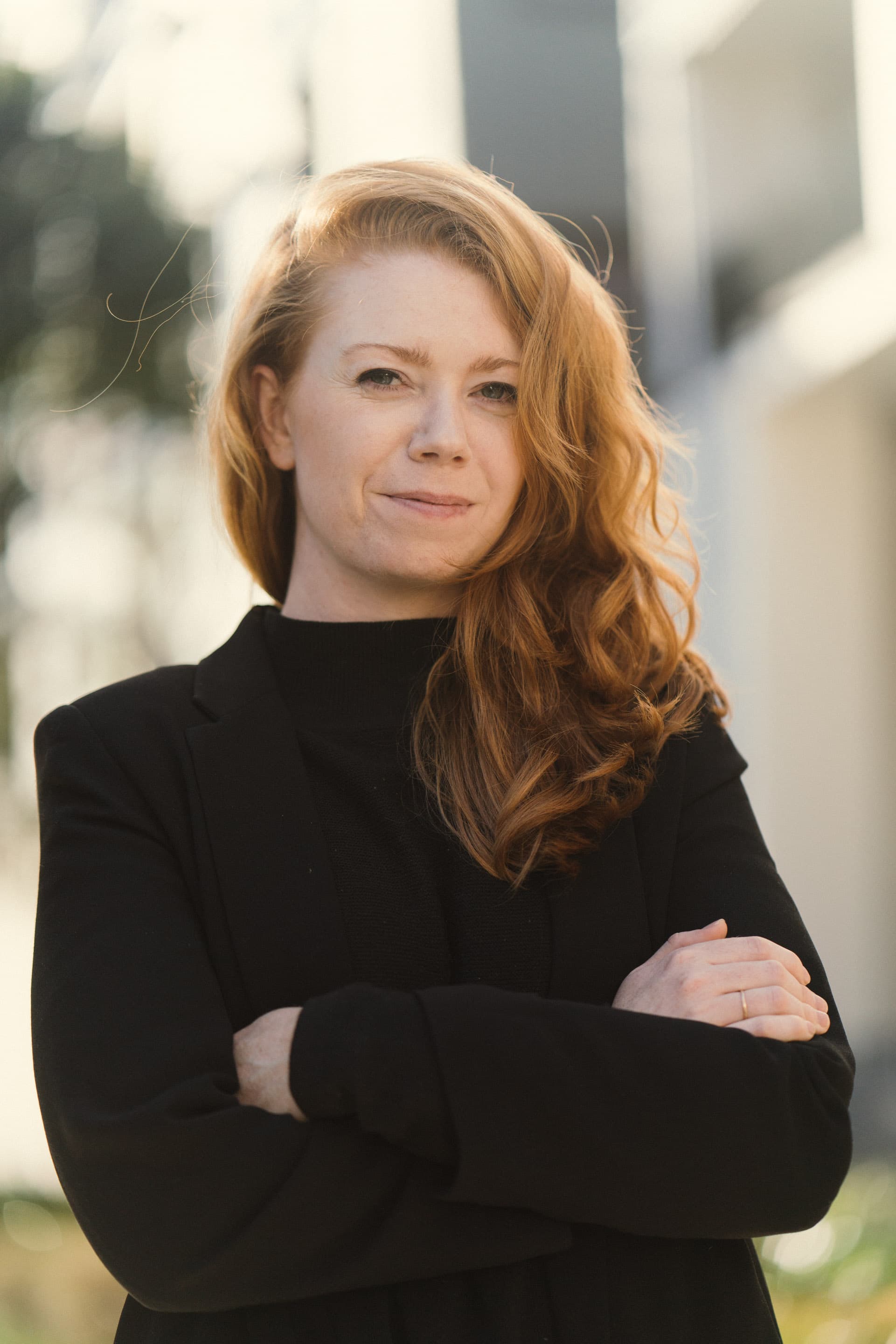 Confident woman with wavy red hair wearing a black blazer, standing with arms crossed.