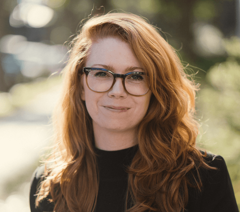 Smiling woman with long wavy red hair and glasses wearing a black top outdoors.